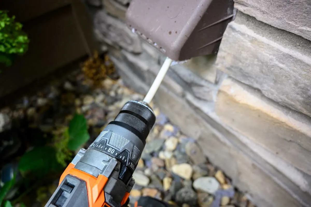 A drill-driven rotary brush fed into an exterior dryer vent hood on a stone-veneer wall